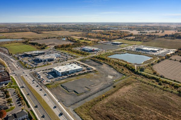 An aerial view of a graded development pad beside a highway, with buildings on one side and open parcels on the other