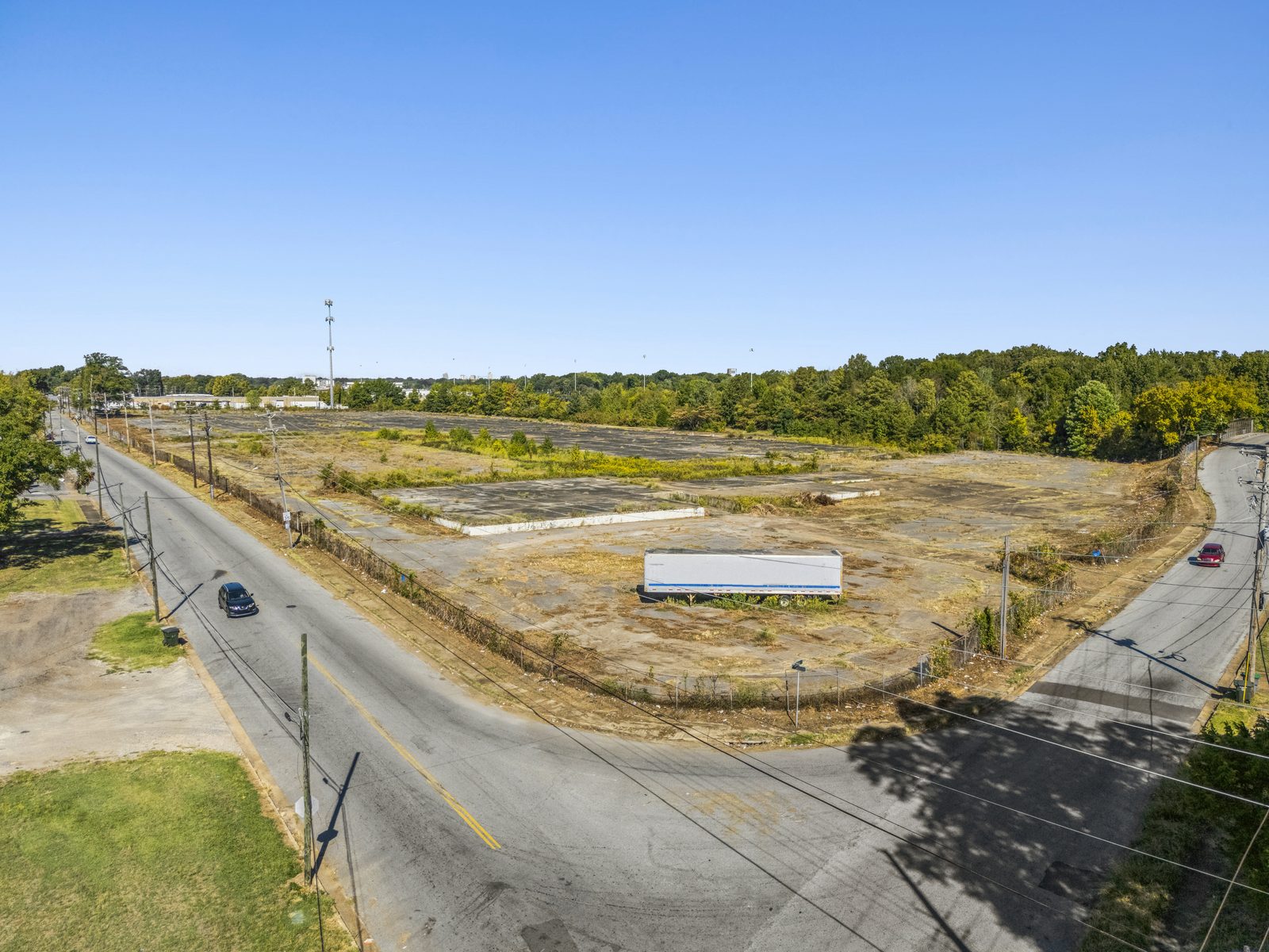 Wide aerial of the site from its corner, with two streets meeting in front of the fenced pad.
