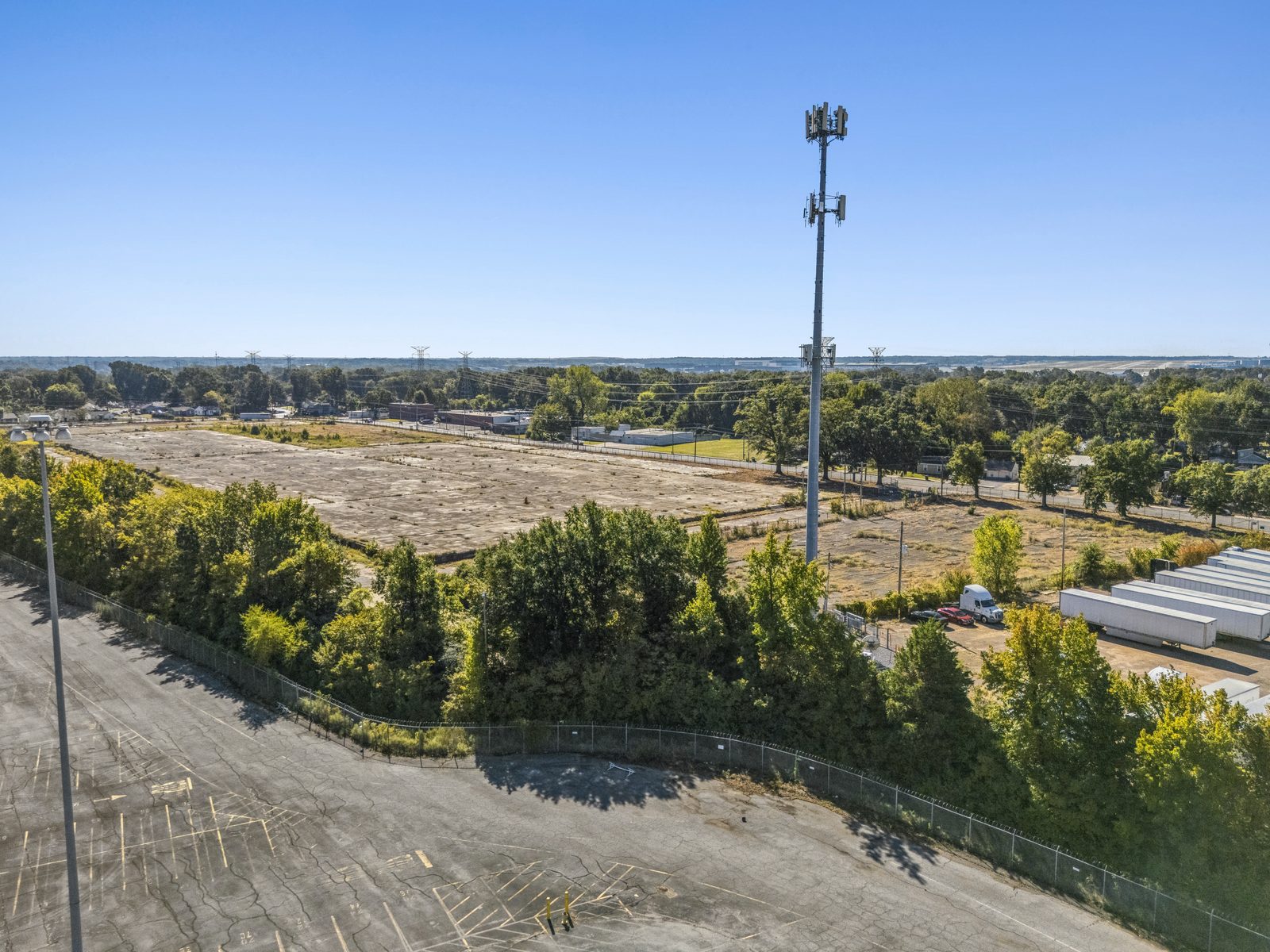 Aerial over the fenced edge of a paved lot, looking past a tree line to the concrete pad.