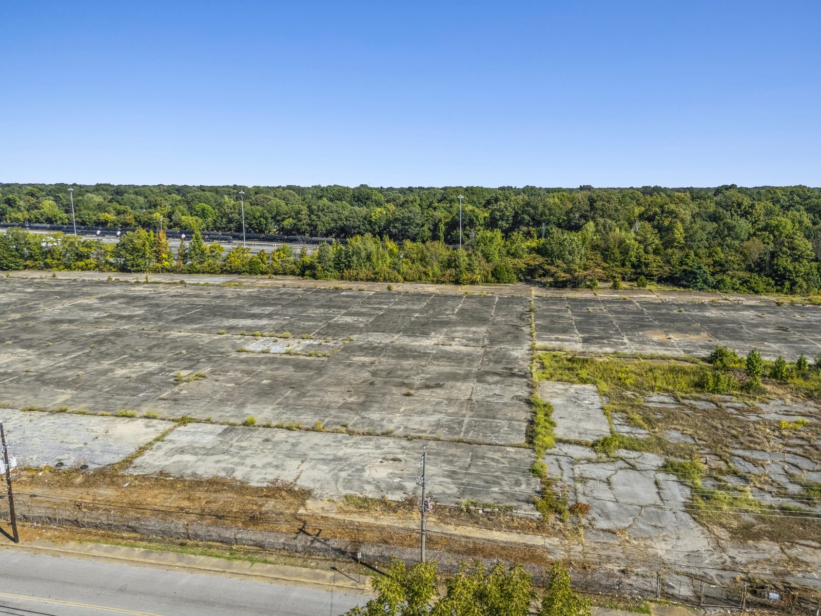 Aerial over the fence and street onto the concrete pad, with a band of trees behind it.