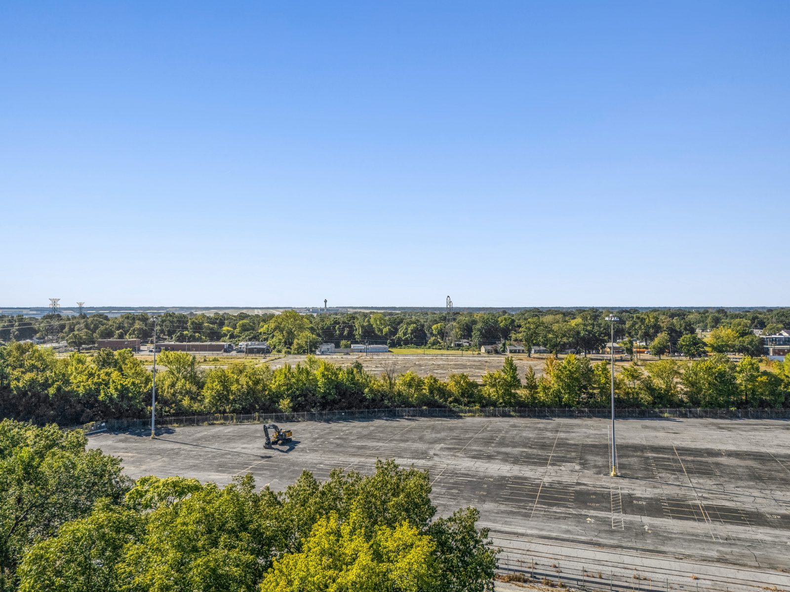 Aerial over a tree line onto the open concrete pad, with a light pole and a piece of equipment on the slab.