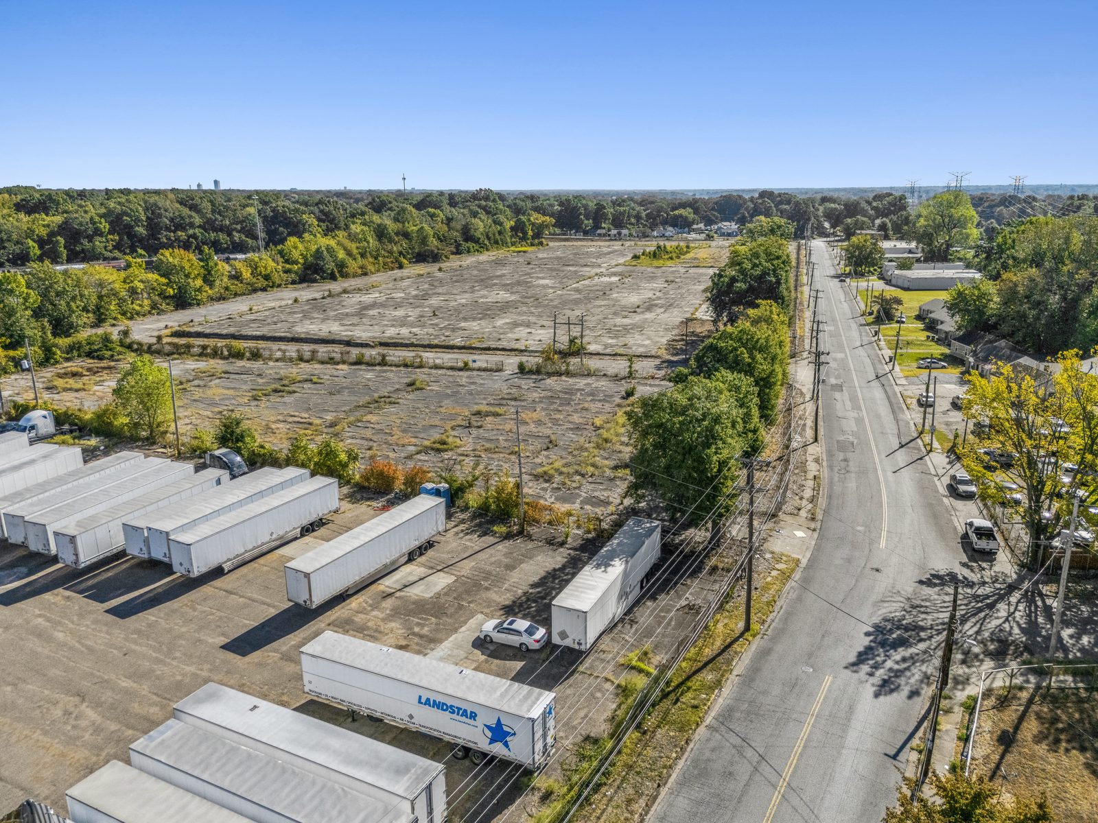 Aerial looking down the street beside the site, with a trailer parking lot in the foreground and the concrete pad behind the trees.