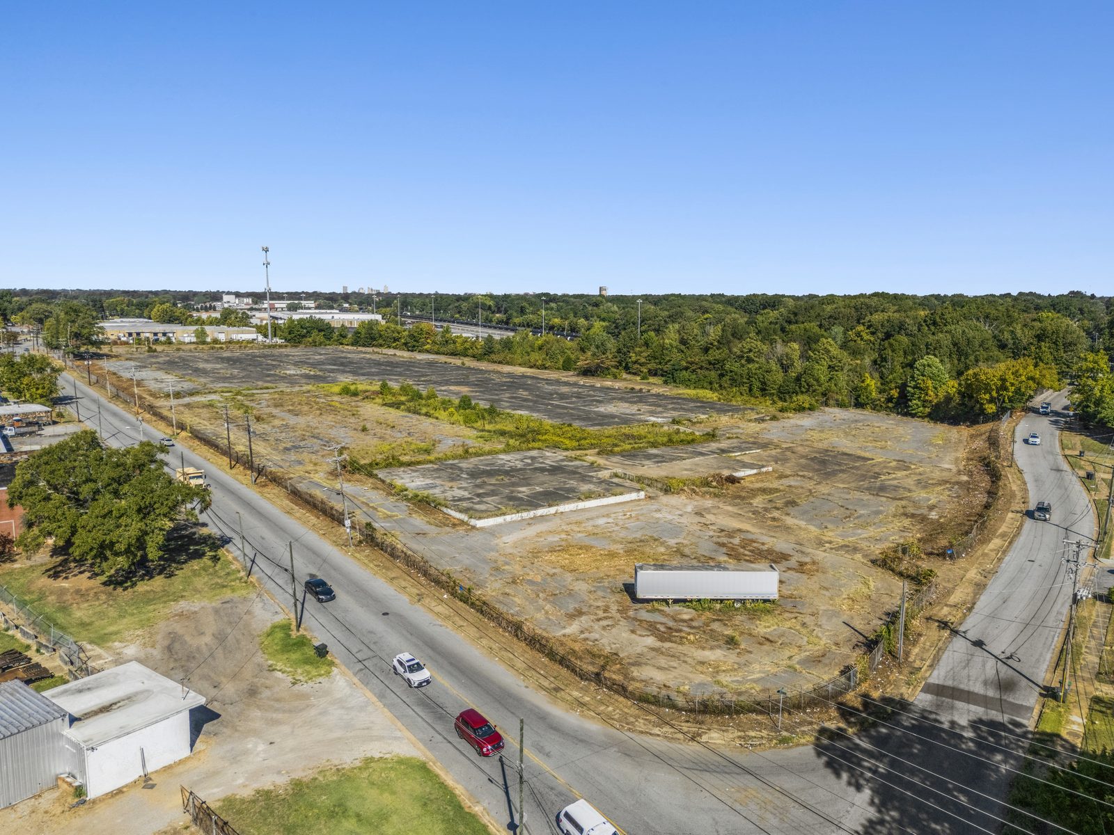 Aerial of the whole site from its corner: the concrete pad inside a fence line, the street curving along two sides, and a small white building at the corner.
