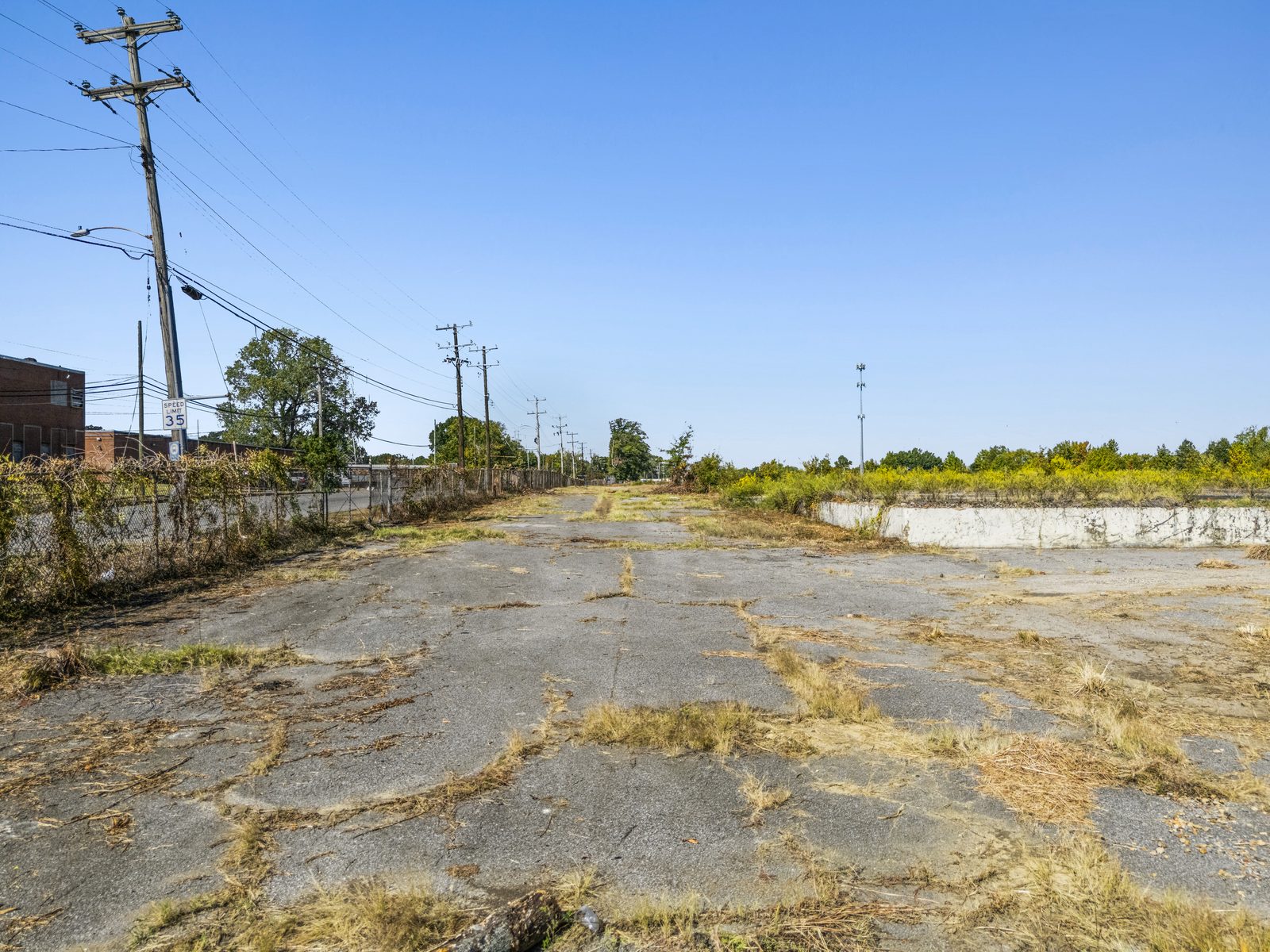 Ground view along the fenced street edge of the site, with power poles and a brick building at left.