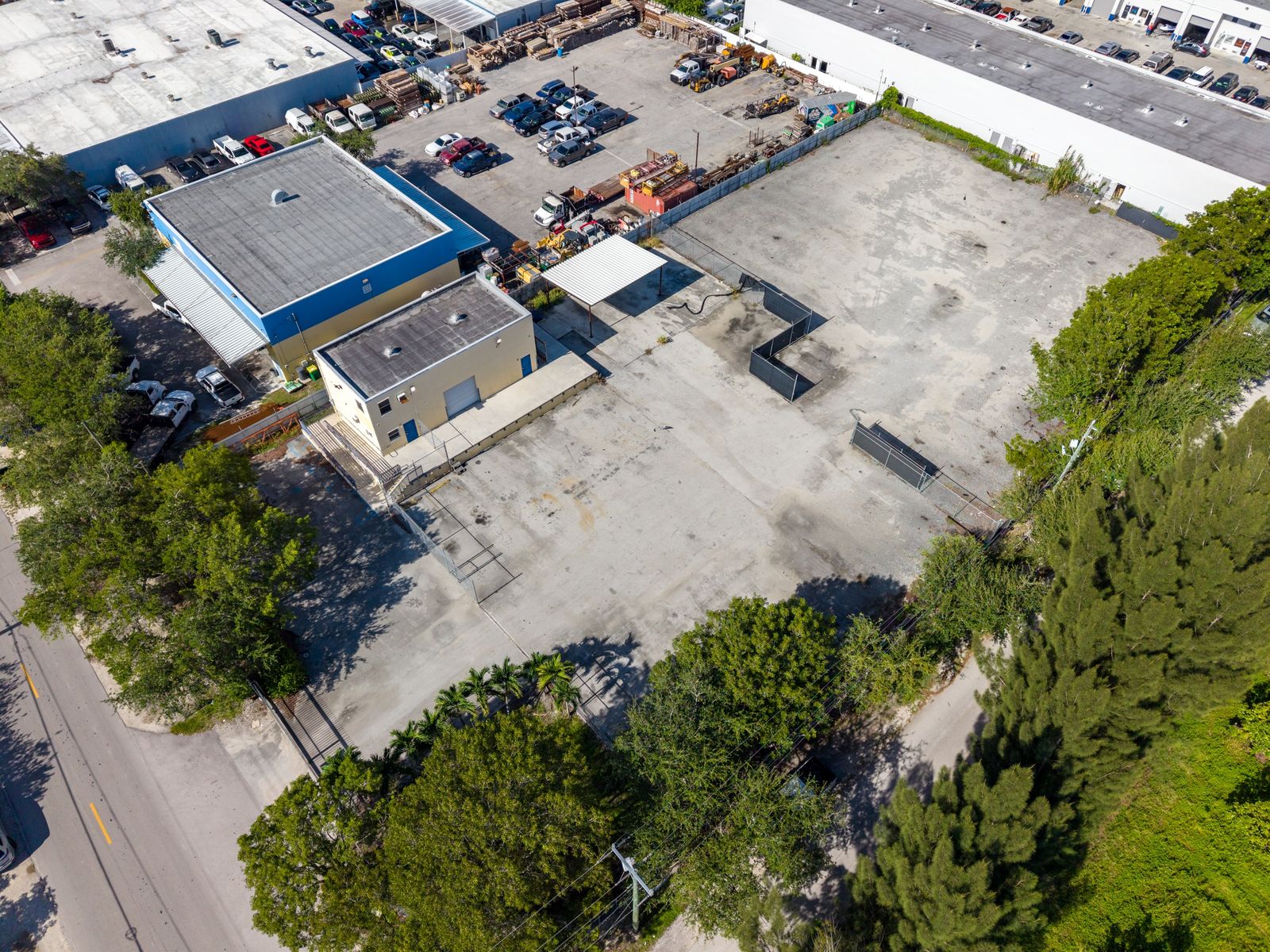 High aerial of the whole site from above the street: the warehouse, loading dock and steel canopy, and the open paved yard behind a tree line.