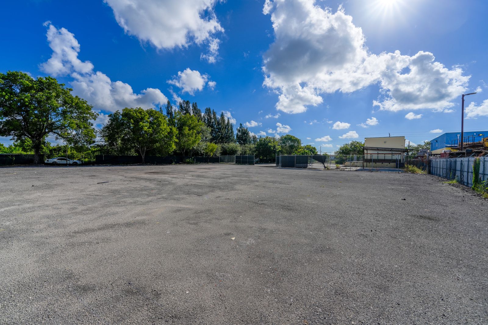 Ground level view across the open paved yard to the fenced tree line, with the warehouse and steel canopy in the distance.