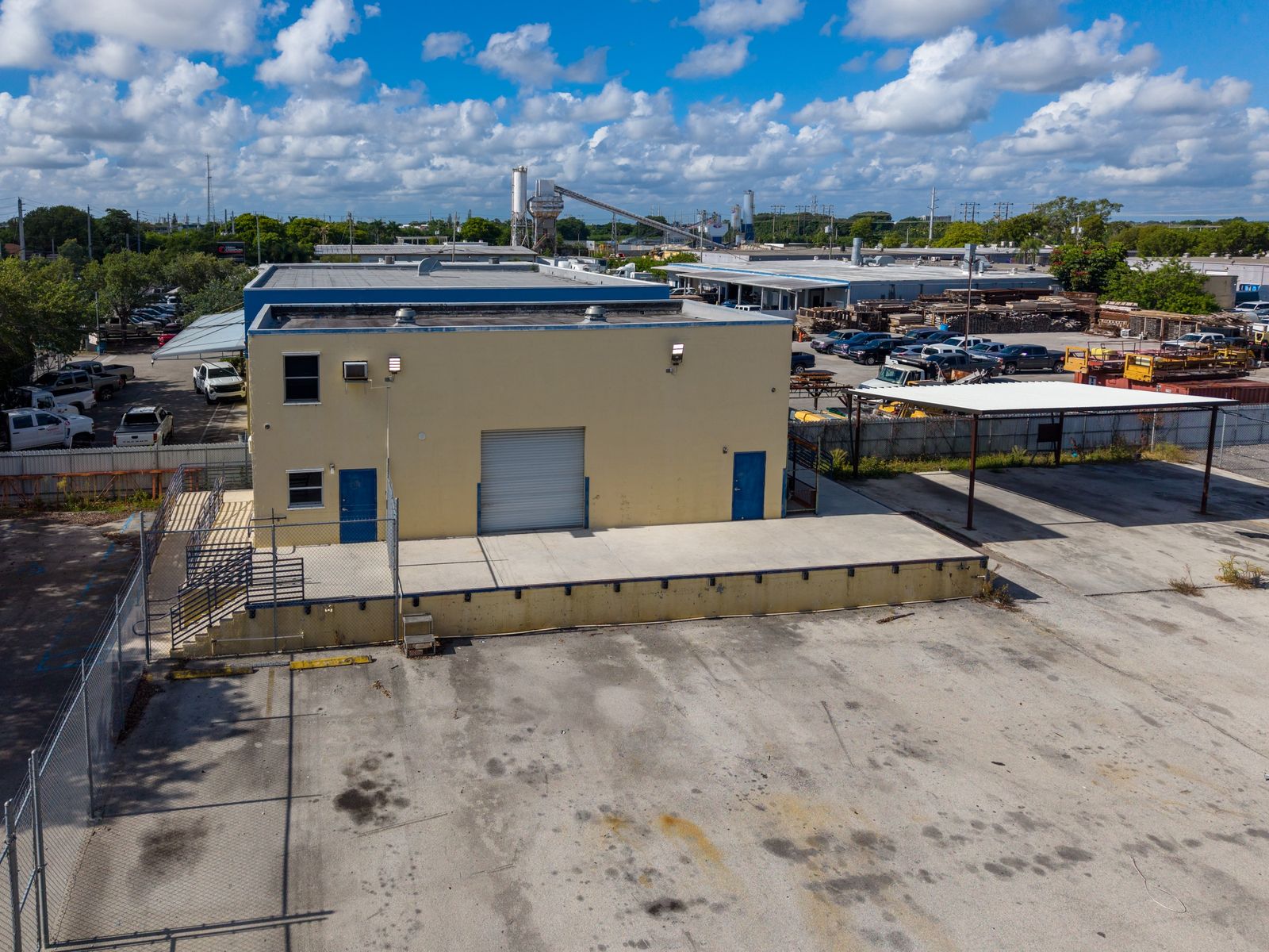 Low aerial of the concrete block warehouse showing the roll up door, the raised loading dock with stairs and ramp, and the steel canopy beside it.