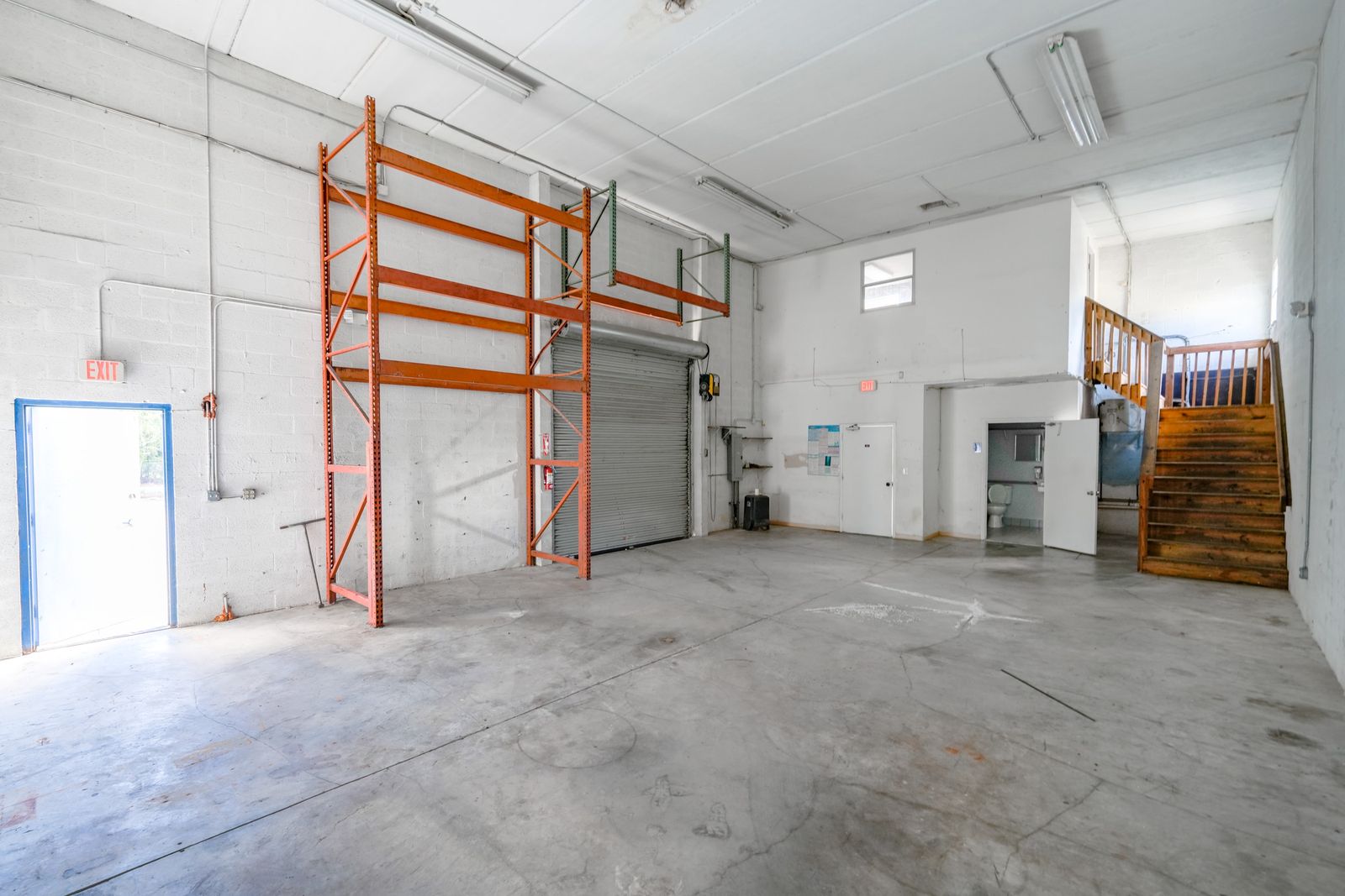 Warehouse interior with pallet racking, the roll up door, a restroom and a wooden stair to the upper level.