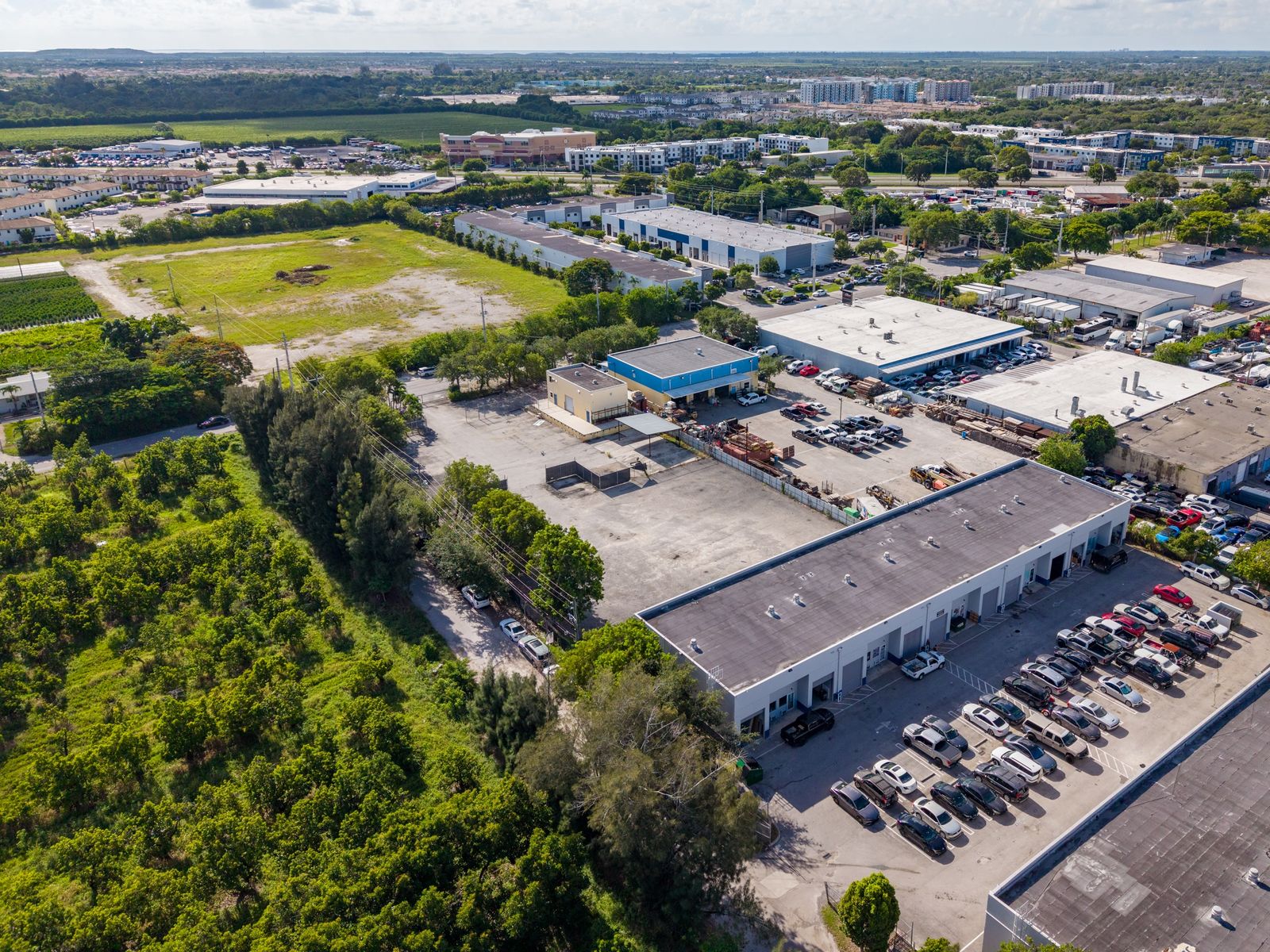 Aerial view from above the site looking across open land toward nearby warehouses and a large parking area.