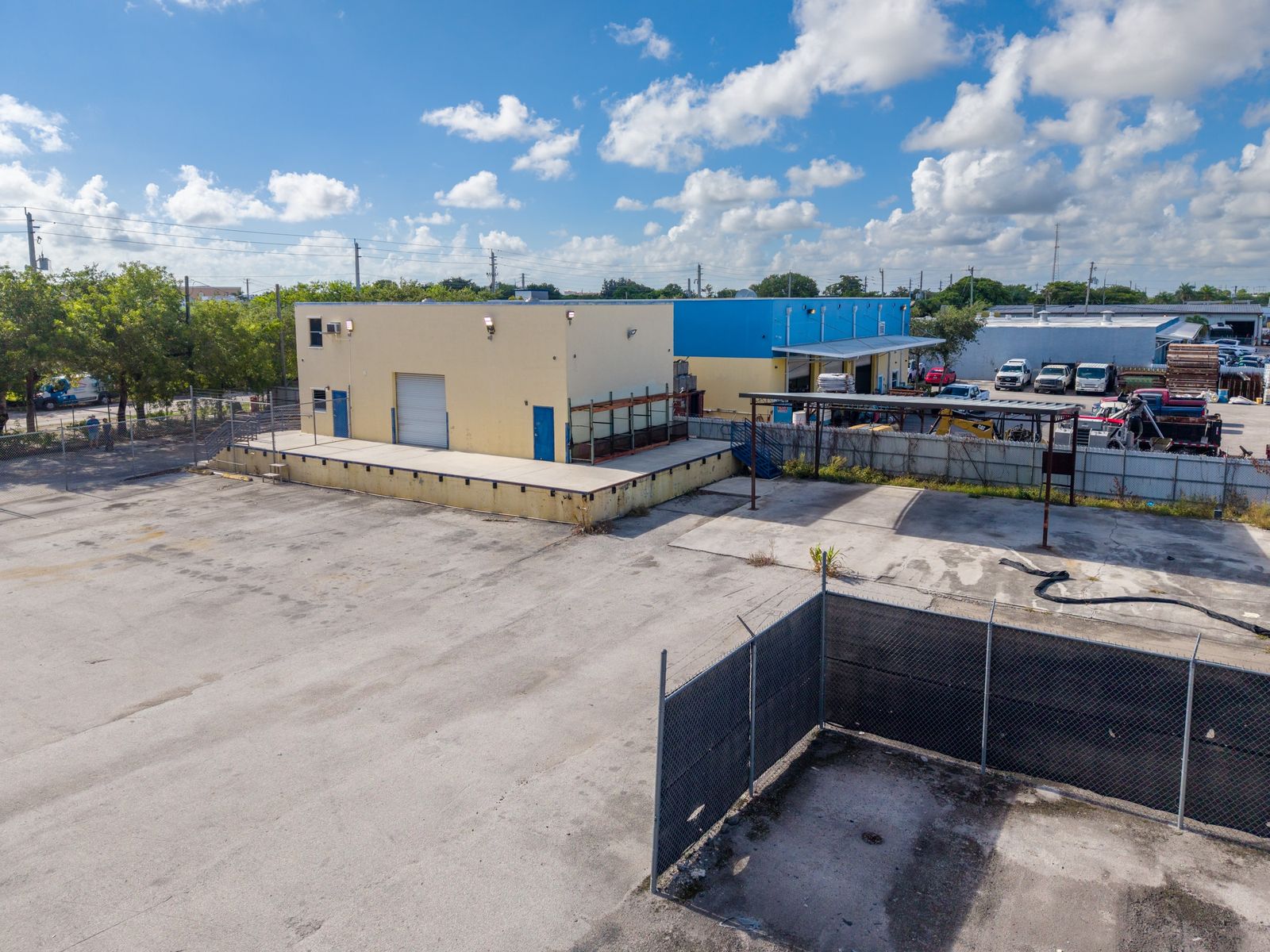 Low aerial of the tan office building and its fenced paved yard, with a canopy structure and trees nearby.
