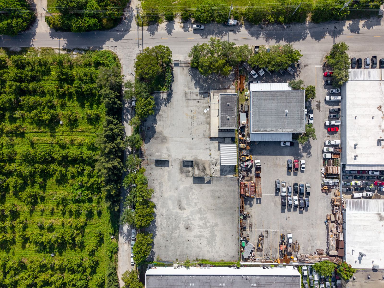 Aerial view of the building and paved lot among neighboring warehouses, parked vehicles and a tree lined street.
