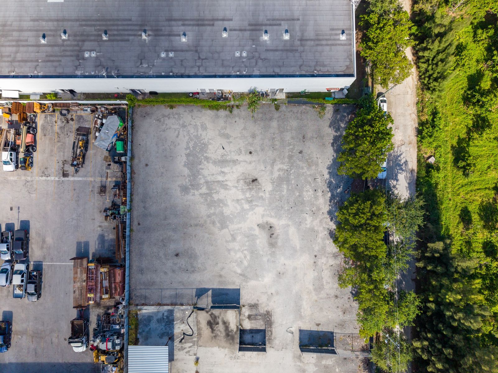 Straight down aerial of the paved yard beside the warehouse roofline, with equipment stored along one edge and trees along the other.