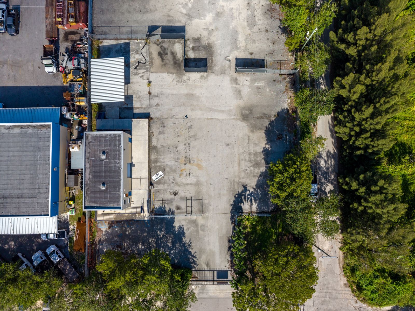 Straight down aerial of the paved yard with a shade structure at one edge and a tree line along the other.