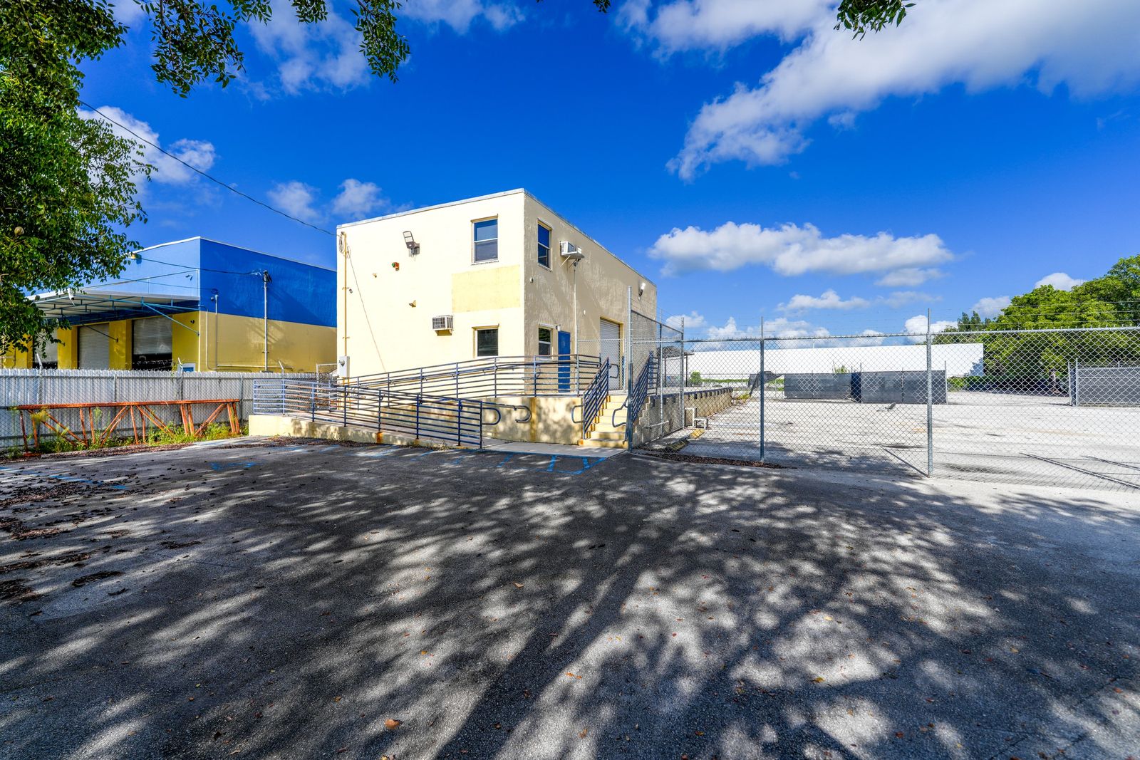 The office building seen from the paved yard, with its entry ramp, railing and the perimeter fence beside it.
