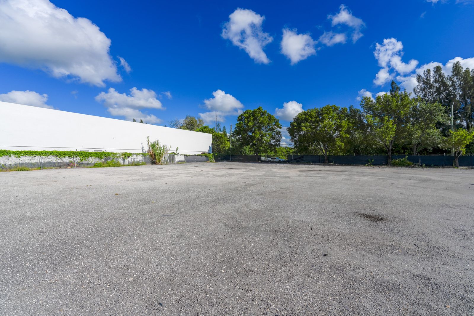 Open paved yard running alongside a long white building wall, with trees along the far edge.